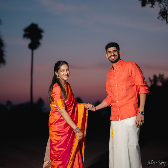A smiling South Indian couple, dressed in vibrant traditional attire, holds hands against a beautiful sunset backdrop with palm trees.
